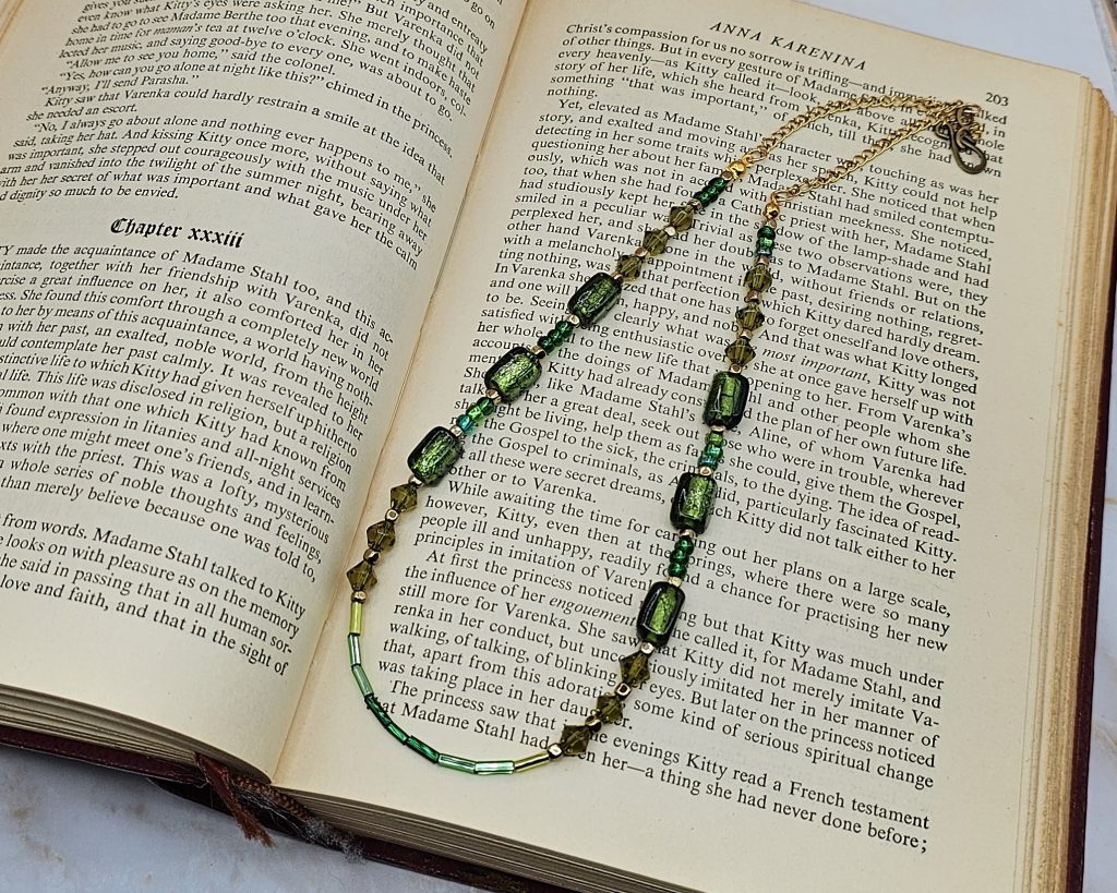 An 18-inch handmade necklace with vintage olive-green glass beads, bicones, and seed beads on a gold-plated stainless steel chain, displayed elegantly on book pages