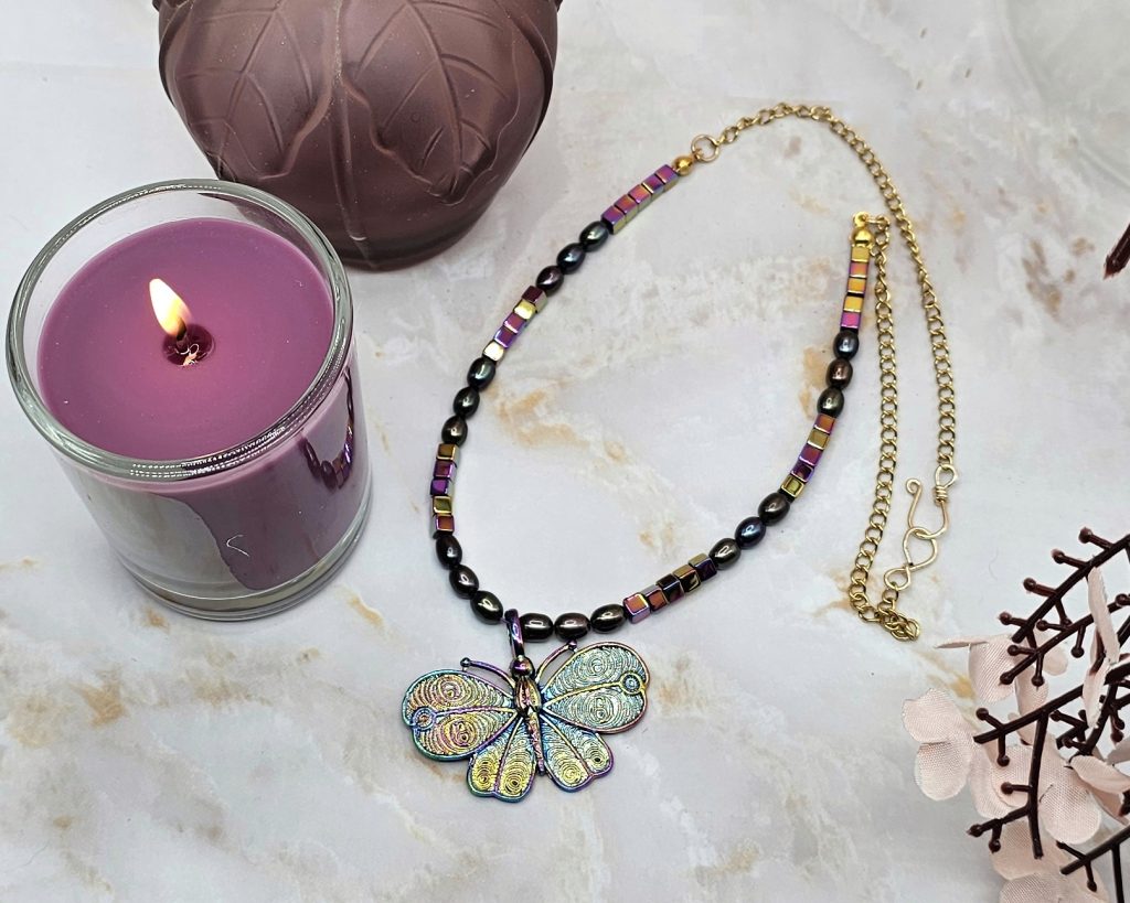 Colorful butterfly pendant necklace with black pearls and rainbow hematite beads on a gold chain displayed on a marbled background with a purple candle.