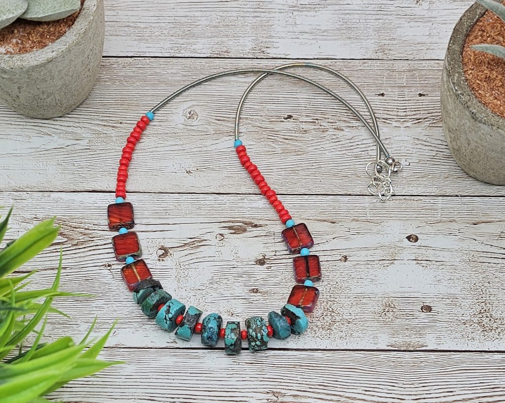 19-20" necklace with rough turquoise rondelle beads and red Czech glass squares, handmade with bold and earthy boho displayed flat on pale wood background with some plants.
