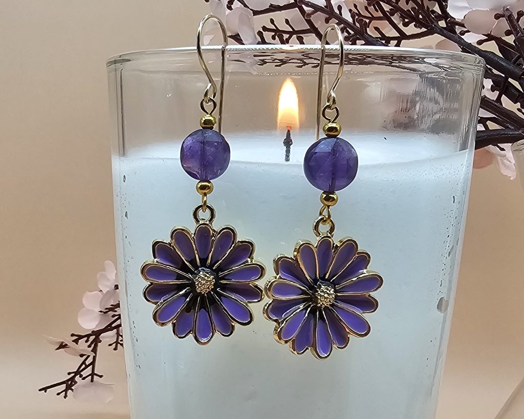 Purple enamel flower earrings with faceted amethyst beads and gold- filled handcrafted ear wires displayed on a candle holder in front of pink flowers and peach background.