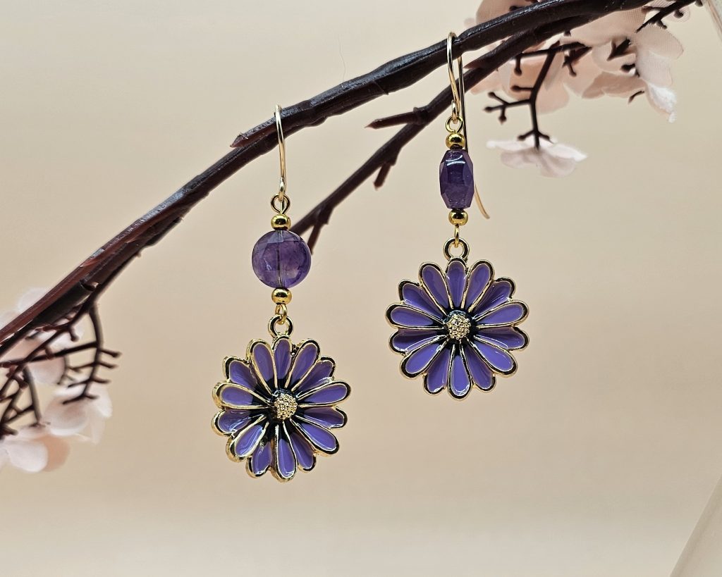 Purple enamel flower earrings with faceted amethyst beads and gold-filled handcrafted ear wires displayed hanging from a flower stem against a beige background.